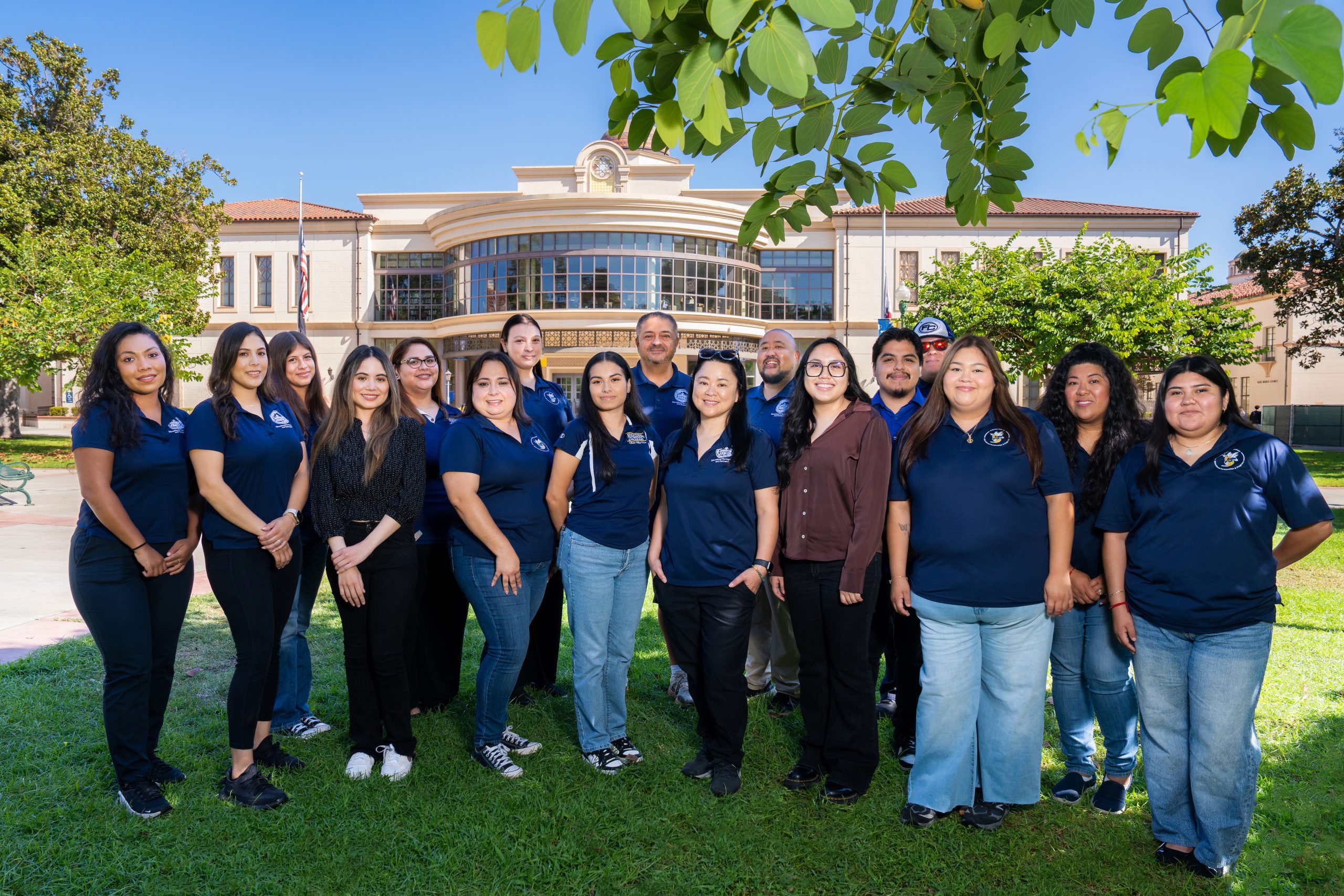 A group of individuals in matching navy blue shirts stands together on grass in front of a large building with a clock tower, under a clear blue sky.