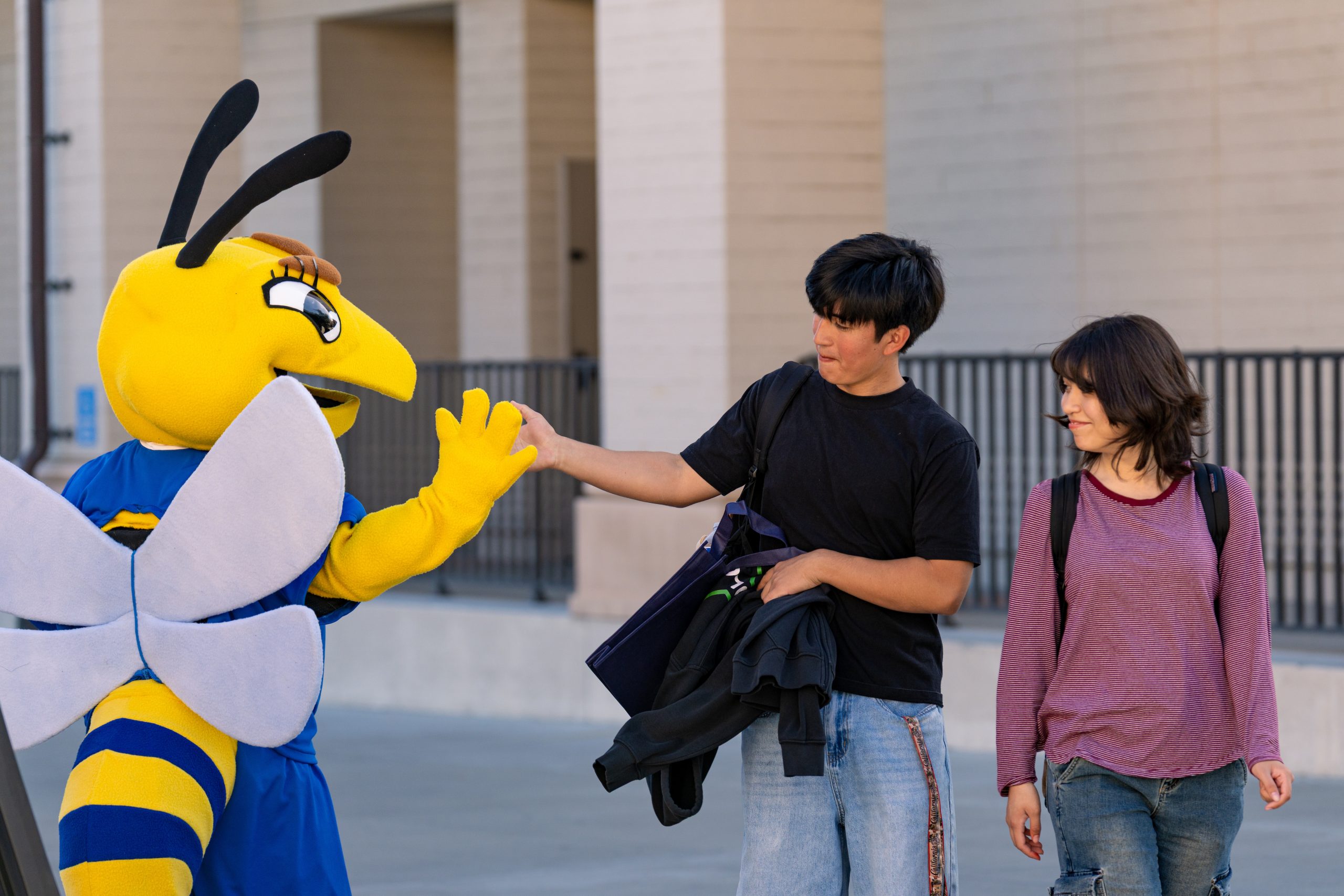 Fullerton College's mascot, Buzzy the Hornet, greets a male student with a high-five while a female student watches the interaction.