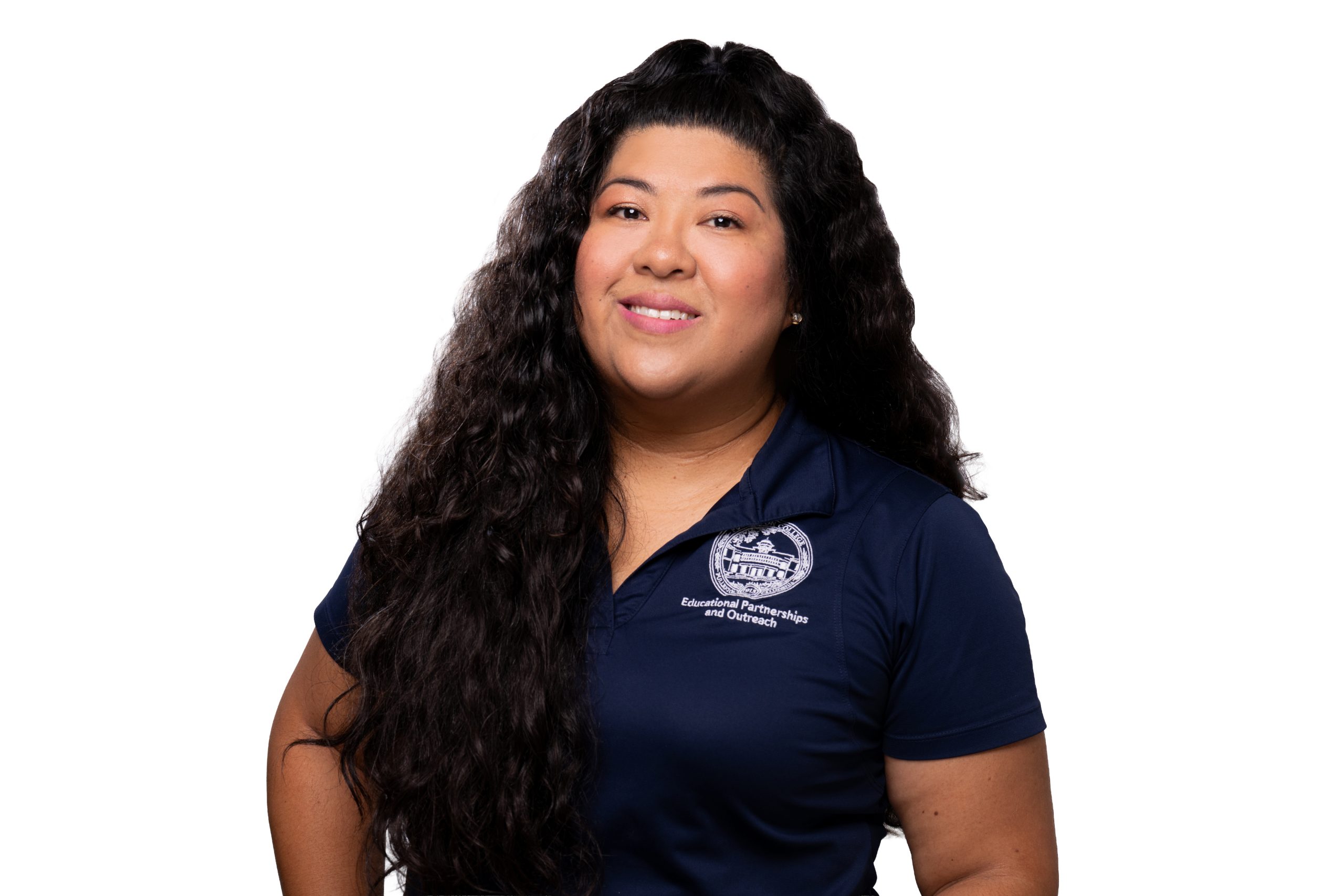 A person with long, dark curly hair smiles while wearing a navy blue polo shirt featuring an embroidered logo and text.