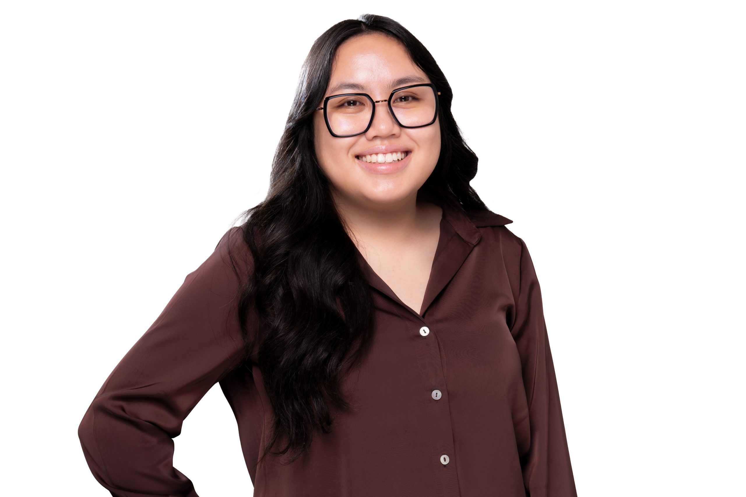 A person with long dark hair and glasses smiles while wearing a brown button-up shirt against a white background.