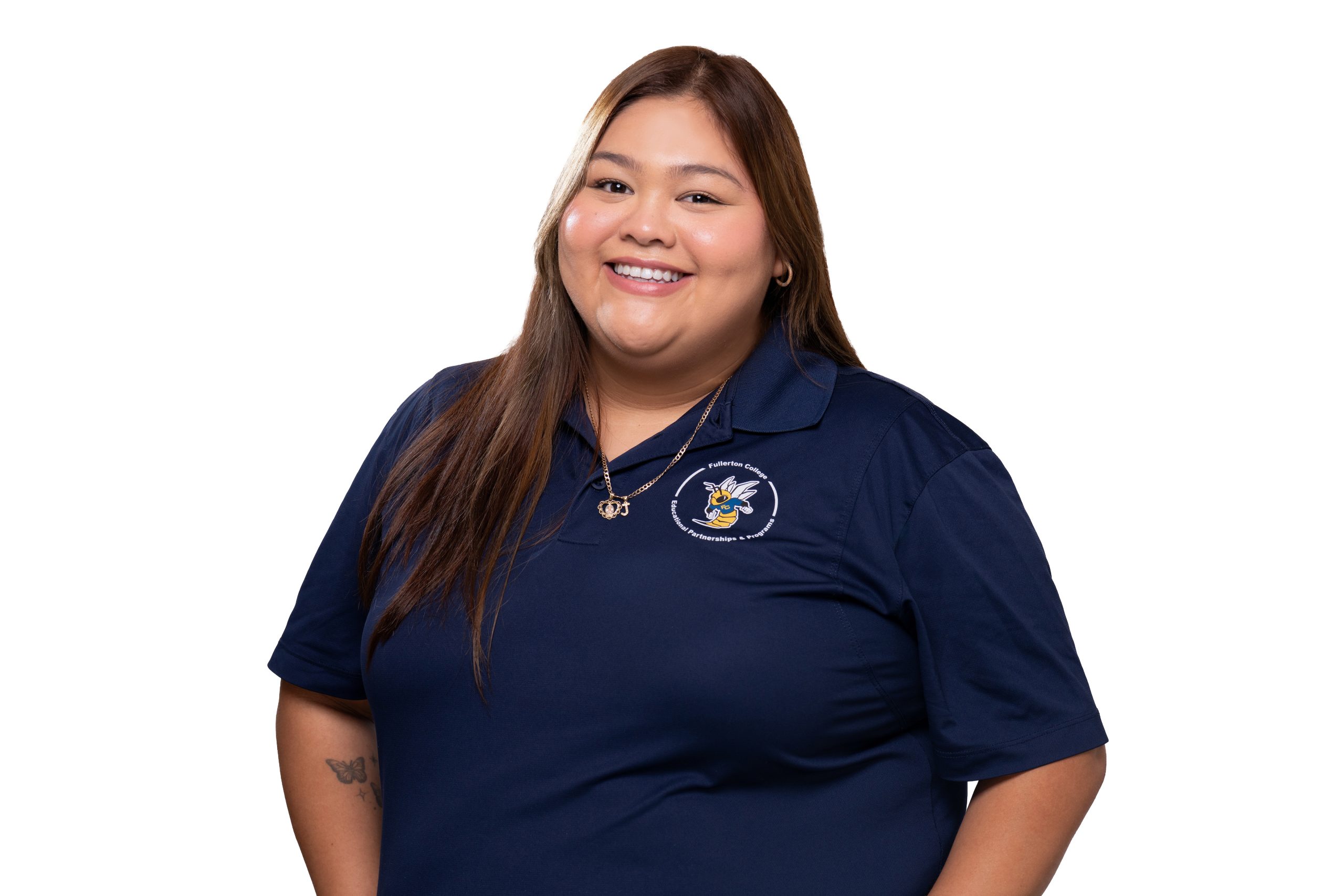 A smiling person with long hair wears a navy blue polo shirt featuring a logo, standing against a plain white background.