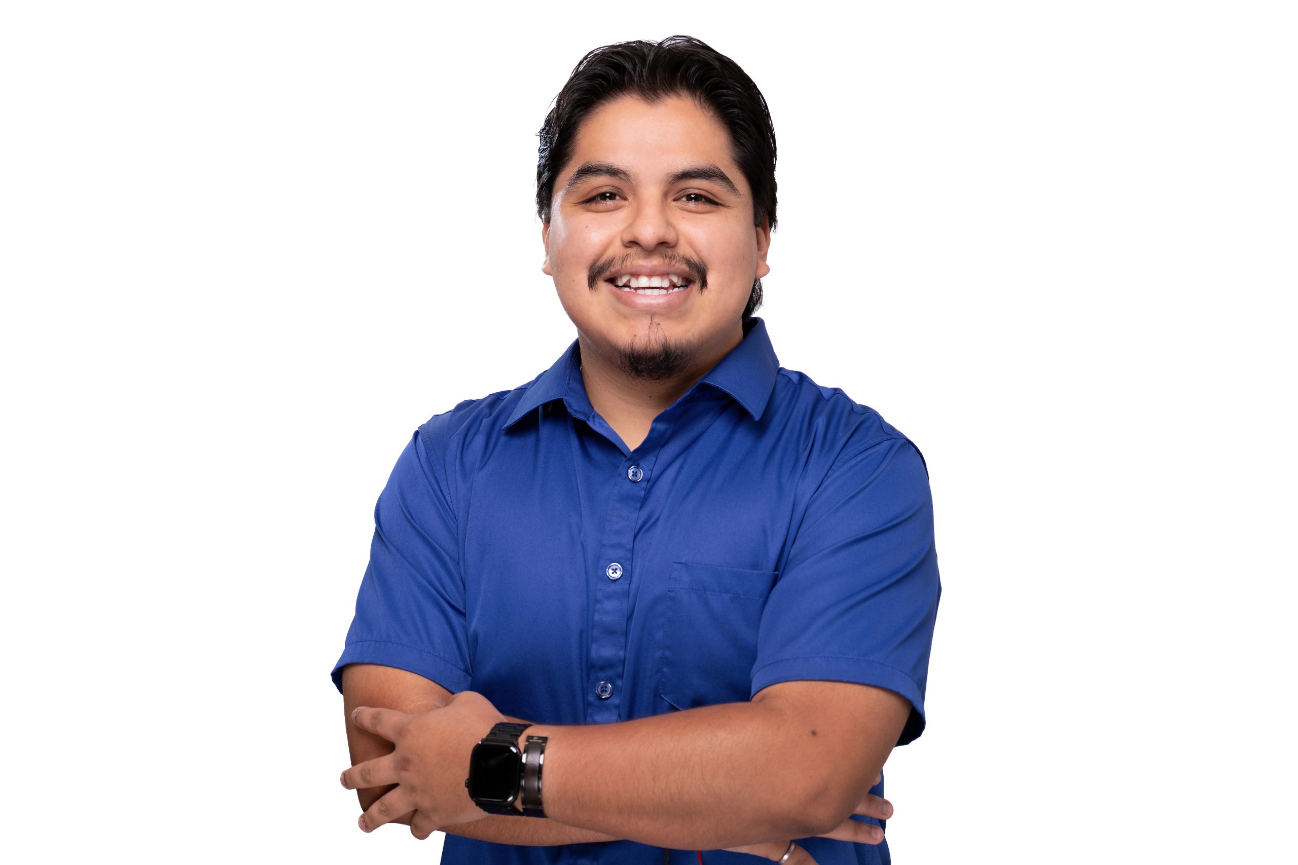 A person with dark hair and a goatee, wearing a blue short-sleeved shirt and a smartwatch, smiles while standing with arms crossed against a white background.