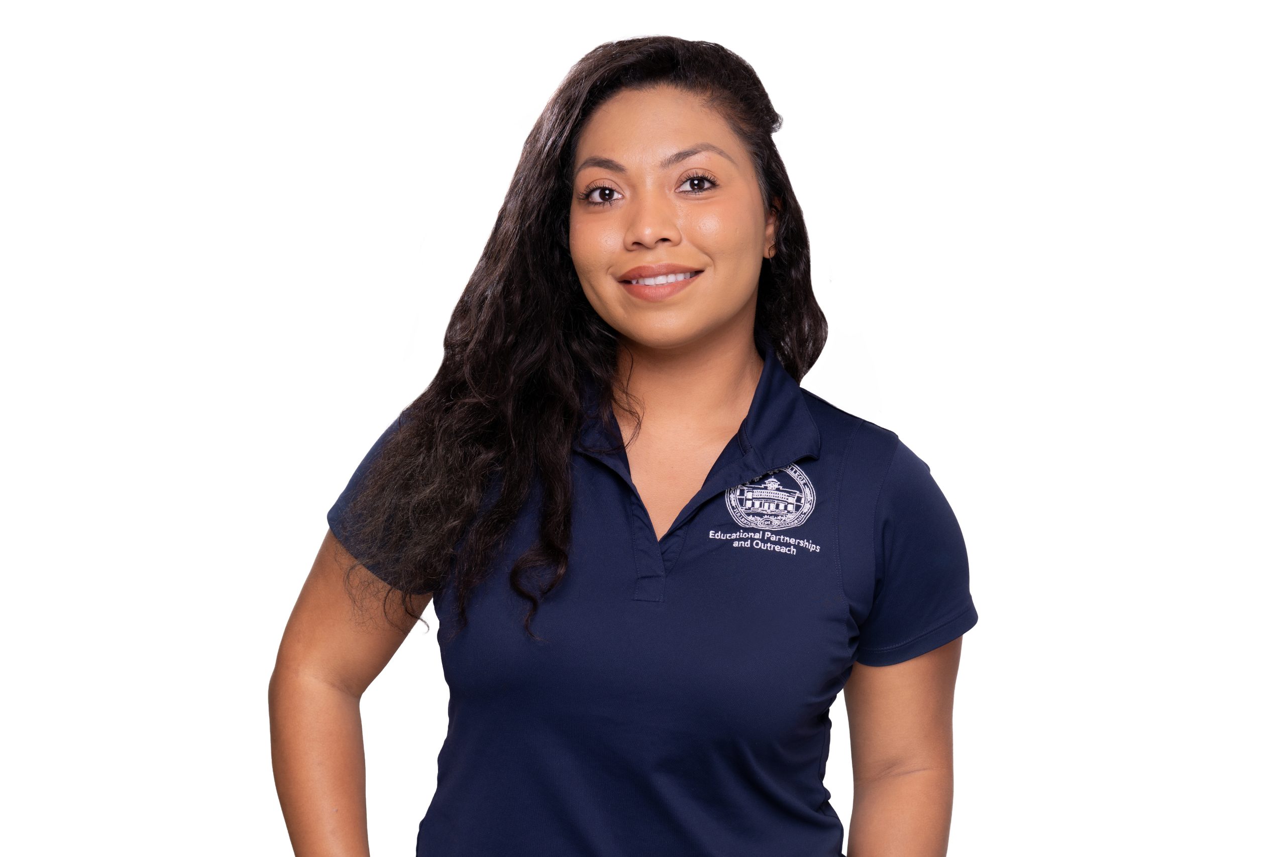 A person with long dark hair smiles while wearing a navy blue polo shirt featuring a logo and text for "Educational Partnerships and Outreach."